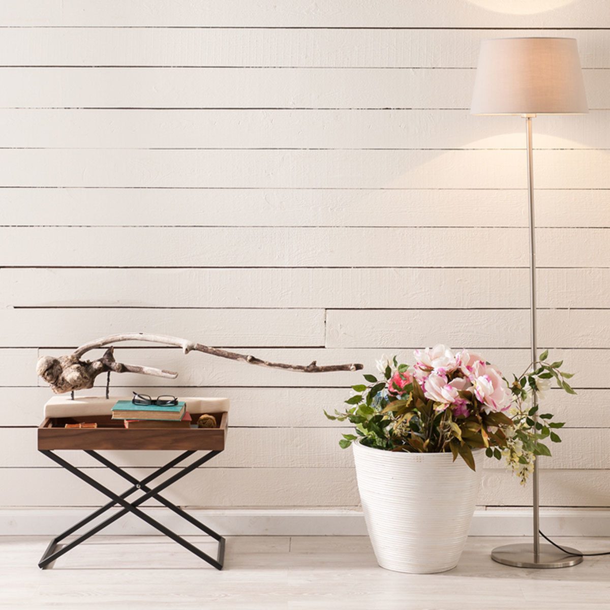A wooden side table holds glasses and a branch, beside a white planter with flowers, near a standing lamp against a light wooden wall.