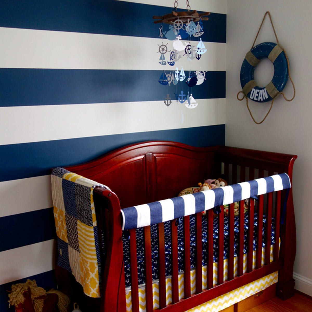 A wooden crib, adorned with a quilt and toys, sits against striped blue and white walls. A mobile hangs above, complementing nautical-themed decor nearby.