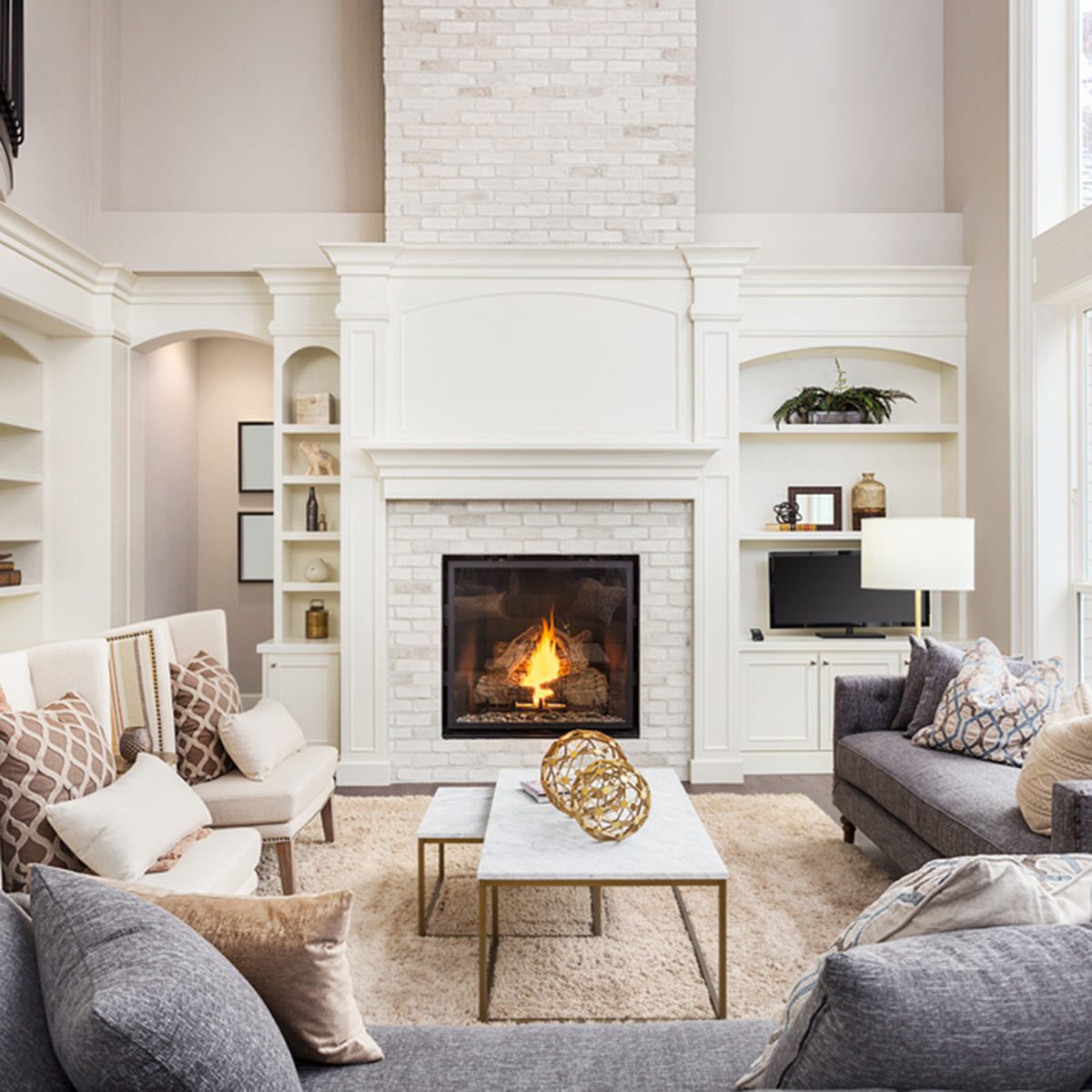 A cozy living room features two seating areas around a marble-topped coffee table, with a fire burning in a white brick fireplace.