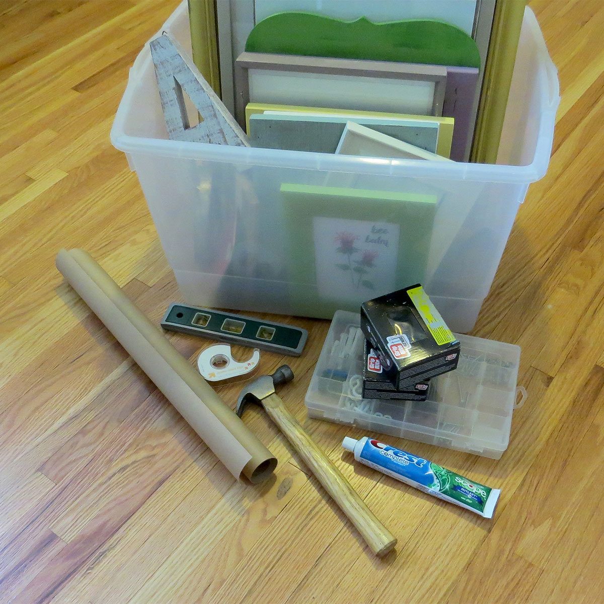 A plastic storage bin holds various picture frames, while nearby lie tools like a hammer, level, and tape, on a wooden floor.