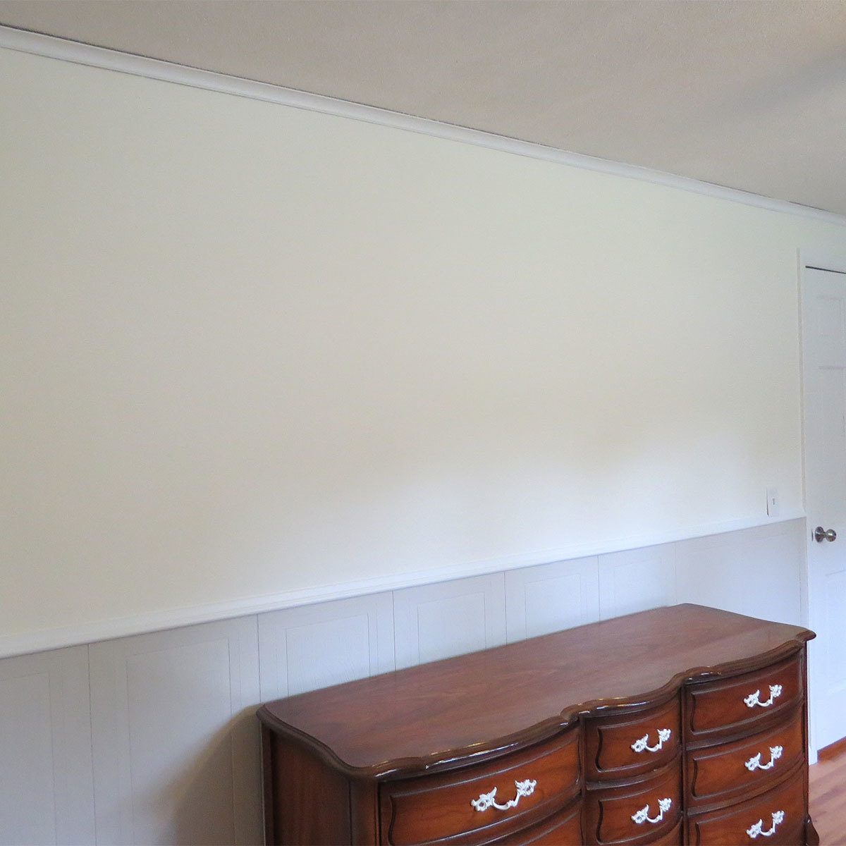 A wooden dresser rests against a light-colored wall in a room with hardwood flooring, featuring a door on the right side of the image.