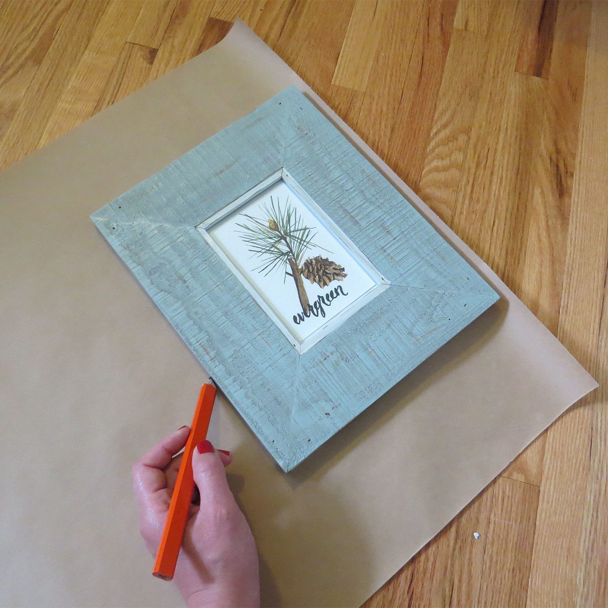 A hand holds an orange pencil, sketching near a wooden picture frame displaying a botanical illustration, placed on a flat surface with paper.