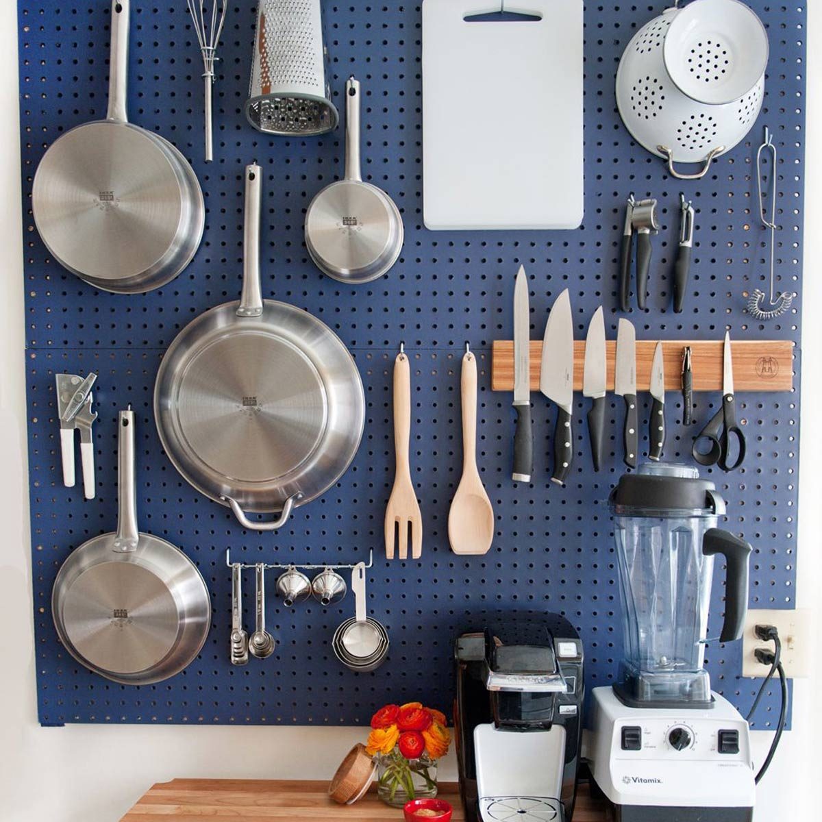 Stainless steel cookware and kitchen tools hang on a blue pegboard, while a countertop blender and coffee maker sit below, accented by a vase of flowers.