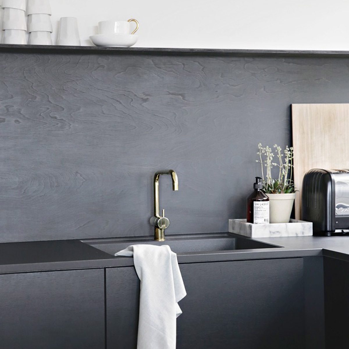 A sleek kitchen sink features a gold faucet, a towel draped over the edge, surrounded by minimal decor and appliances on a dark surface.
