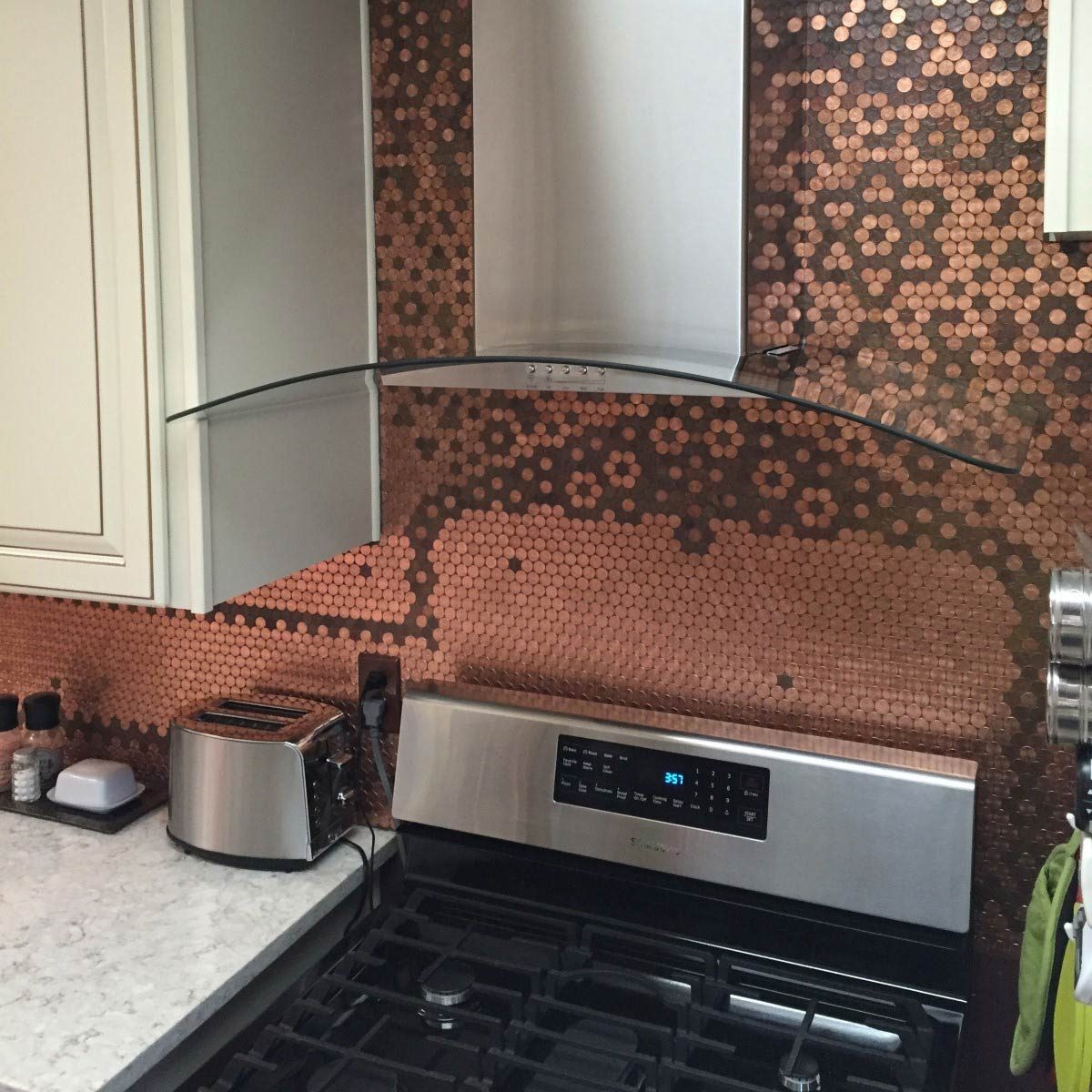 A stainless steel range hood hovers above a gas stove, reflecting light, while a copper hexagon backsplash adds texture in a modern kitchen.