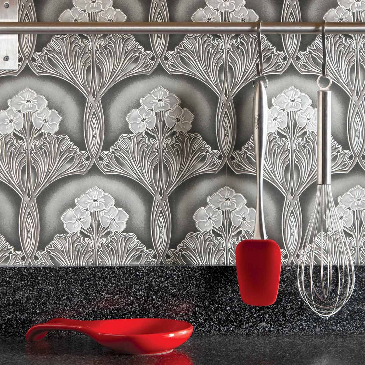 A red spatula and whisk hang from a metal rod against a patterned wallpaper, with a red spoon resting on a black granite countertop.