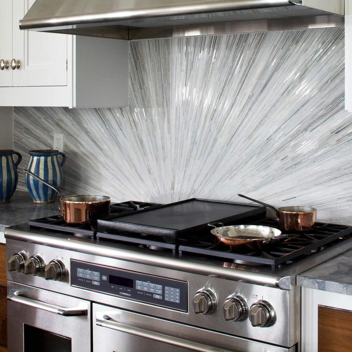 A stainless steel stove features multiple pots, with one resting on a griddle. A decorative tiled backsplash radiates behind, enhancing the kitchen setting.