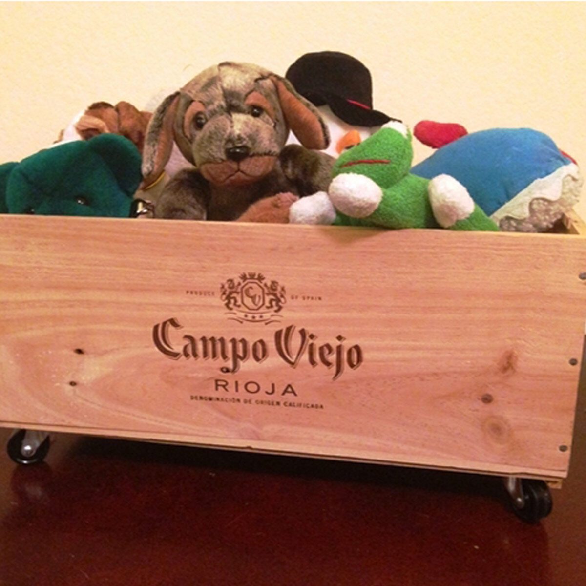 A wooden box labeled "Campo Viejo" holds various colorful stuffed animals, partially peeking out. The box sits on a surface that reflects light.