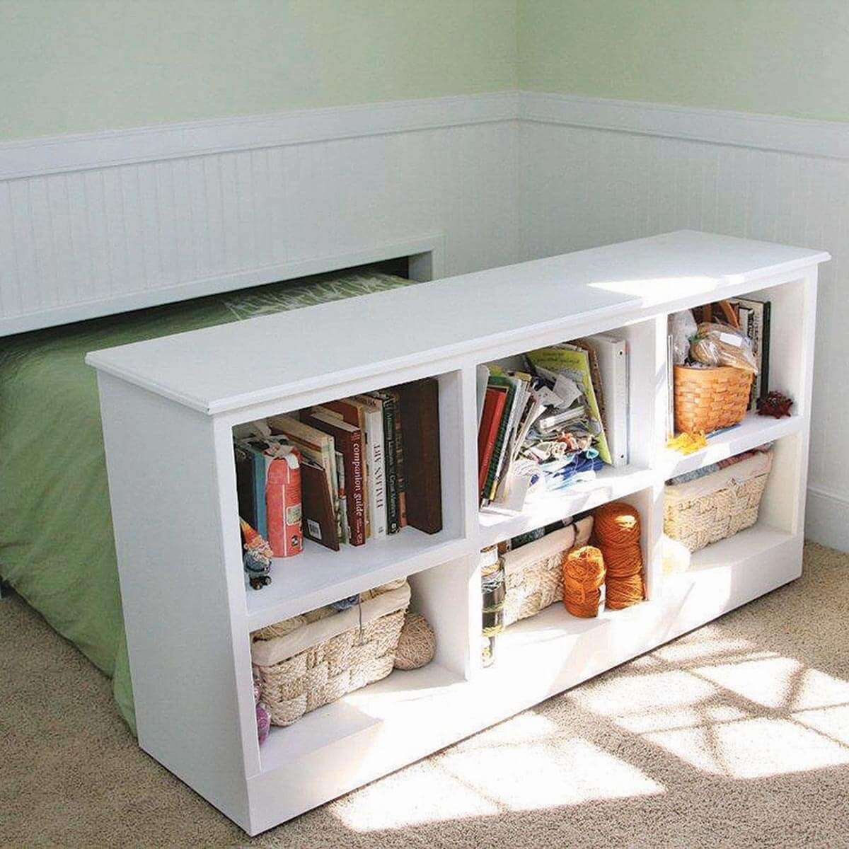 A white bookshelf holds various books and baskets, situated in a sunlit room with light green walls and carpet, next to a bed covered in green bedding.