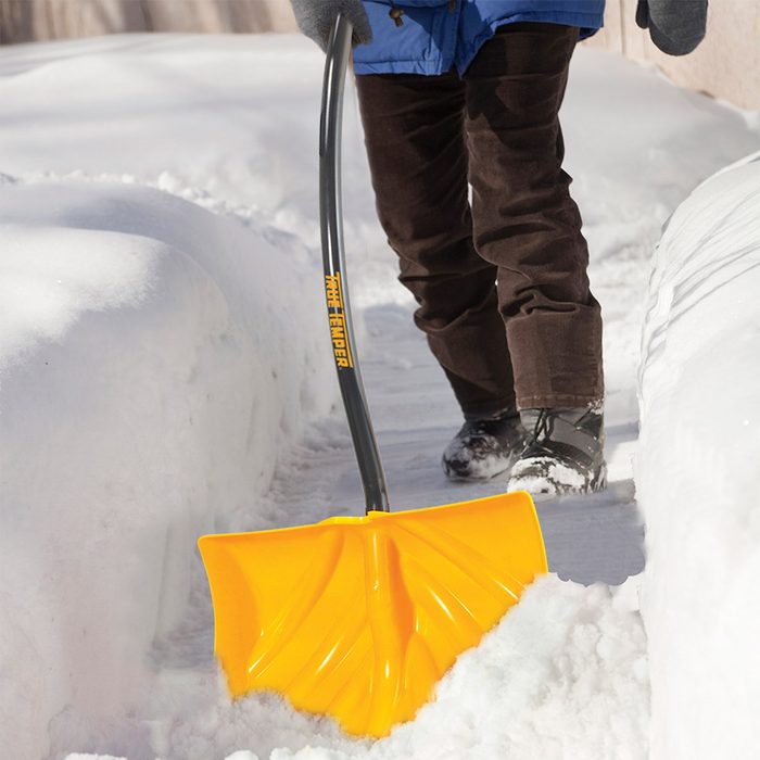A person shovels snow with a yellow snow shovel, creating a clear pathway through a snowy outdoors, while wearing warm clothing and boots.