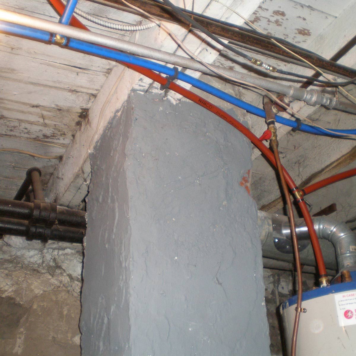 A gray concrete pillar stands amid exposed pipes and wires in a dimly lit basement area, supporting the overhead structure.