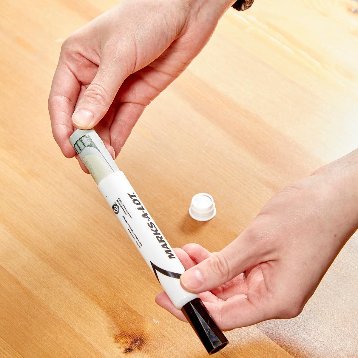 A hand holds a marker while removing a cap; a rolled-up bill is inside the marker, resting on a wooden surface.