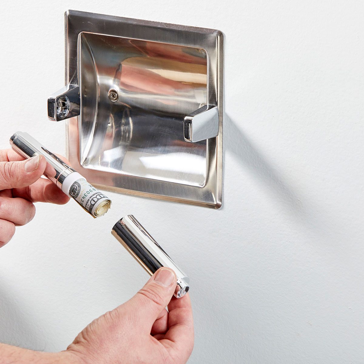 A hand holds a silver tube, partially unscrewed, near a wall-mounted metal receptacle. The clean, white wall serves as the context for this action.