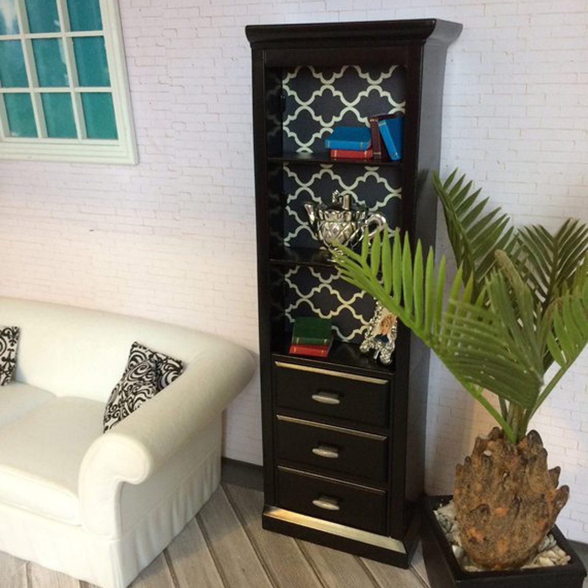 A black bookshelf displays books and decor while a white couch and potted plant complement the modern, cozy room with a light brick wall.