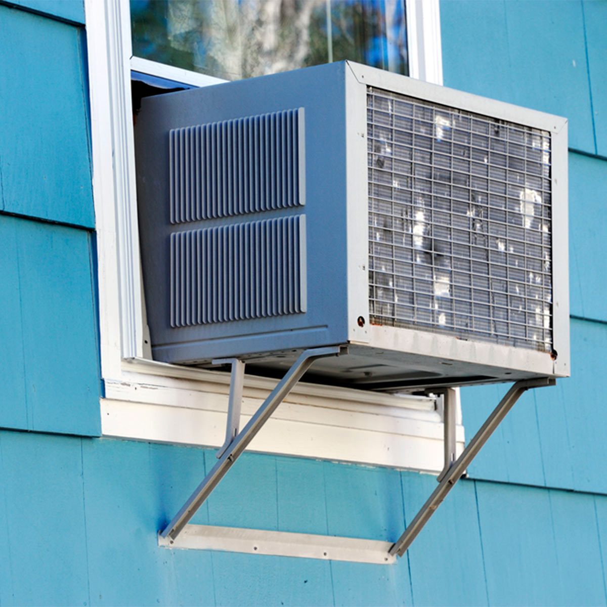 An air conditioning unit is installed through a window, cooling the interior of a blue-paneled building while extending outward for airflow.
