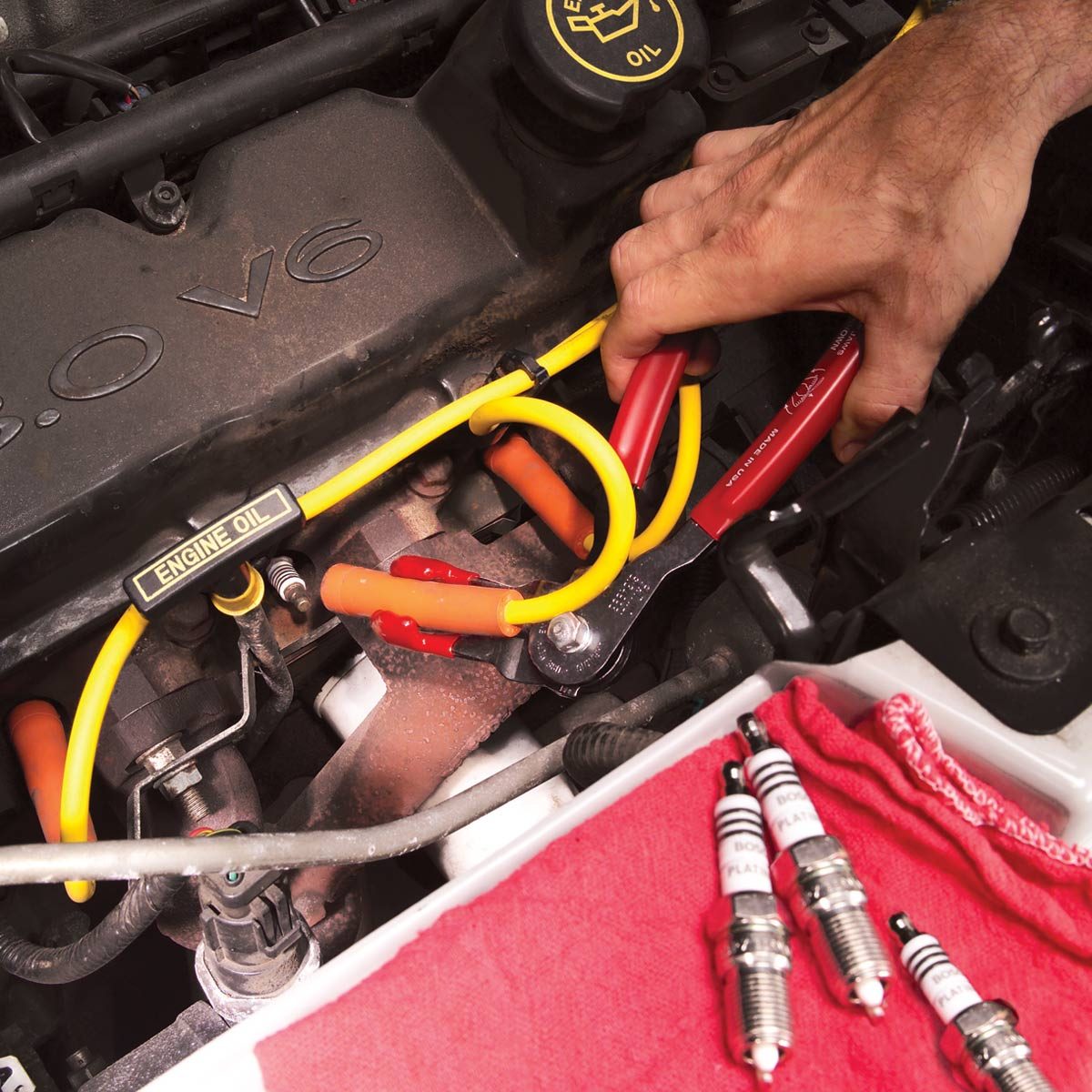 A hand uses a red tool on a car engine, surrounded by wires, spark plugs, and a red cloth, indicating maintenance work in progress.