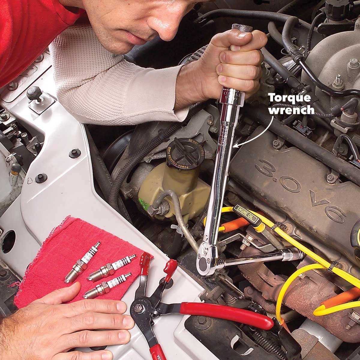 A mechanic uses a torque wrench to tighten bolts in an engine compartment, surrounded by tools and spark plugs on a red cloth.