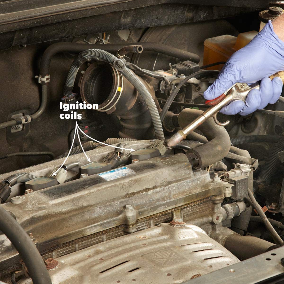 A gloved hand uses a tool to connect or disconnect ignition coils on an engine, surrounded by various engine components and wiring in a vehicle.