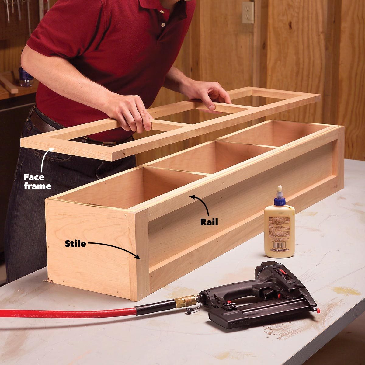 A person assembles a wooden face frame on a box structure, with glue and a nail gun nearby, in a workshop setting.