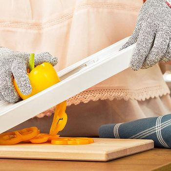 A hand holds a yellow bell pepper, slicing it with a mandoline slicer. A wooden cutting board and kitchen towel are in a bright, cozy kitchen setting.