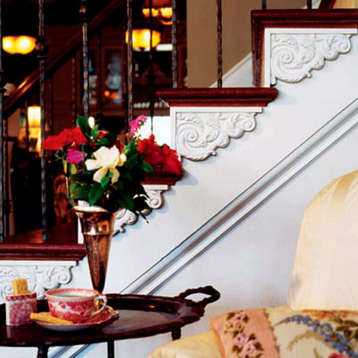 A decorative vase holds colorful flowers, resting on a side table with a cup and cookies, beside a staircase featuring ornate woodwork and a cozy armchair.