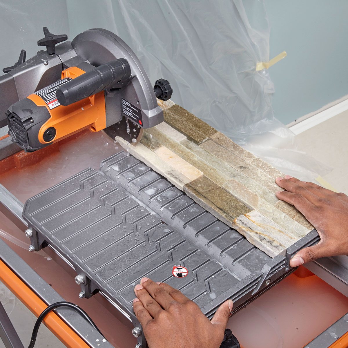 A person uses a wet saw to cut stone tiles on a workstation, surrounded by plastic sheeting and a light-colored wall. Water splashes from the saw.