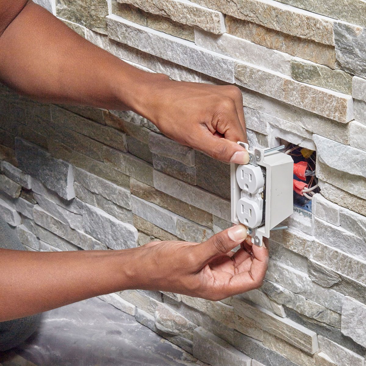 A person is installing an electrical outlet into a wall made of stone tiles, working with exposed wiring in the background.