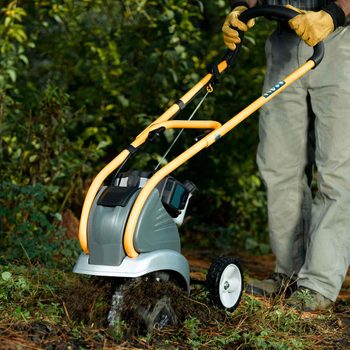 A garden tiller is being pushed through soil, turning over dirt and debris in a lush, green area surrounded by trees and foliage.