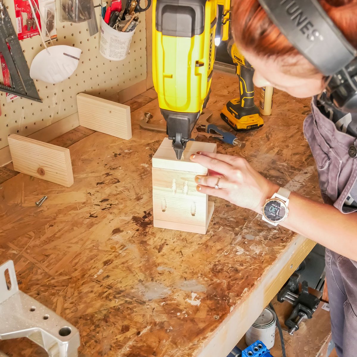 A person uses a nail gun to attach wooden pieces on a workbench amidst various tools and materials in a workshop environment.