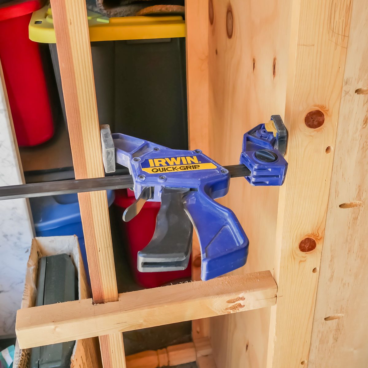 A blue clamp is securing two wooden pieces together, positioned against a light-colored wood wall, with storage containers visible in the background.