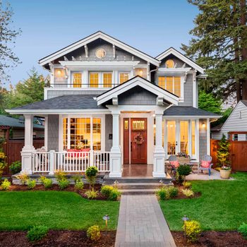 A modern gray house with decorative trim stands before a manicured lawn, featuring a welcoming porch and warm lights glowing from within.