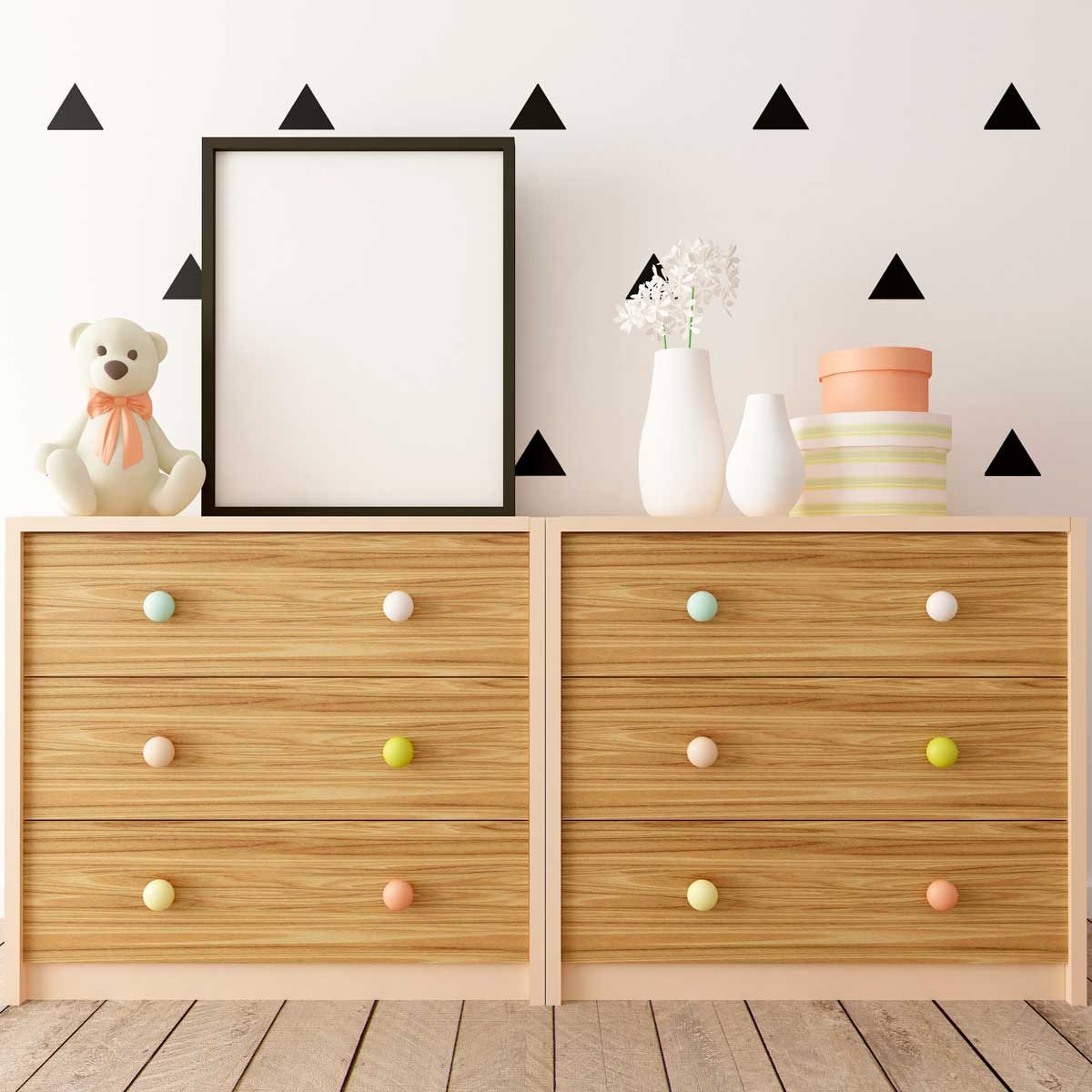 Two wooden dressers with colorful knobs hold a teddy bear, vases, and decorated boxes, against a wall featuring black triangle decals and a blank frame.