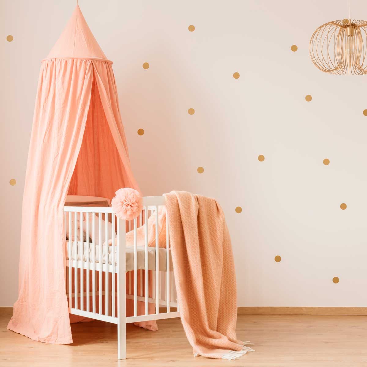 A white crib with a soft pink blanket is beside a coral canopy, against a light wall adorned with gold dots, creating a cozy nursery atmosphere.