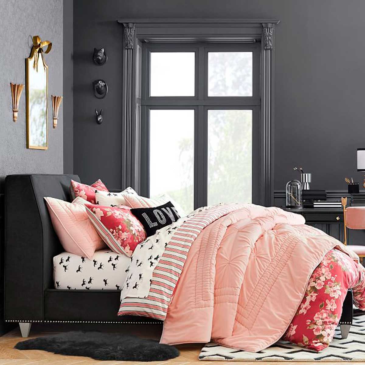 A styled bed with multiple patterned pillows and a pink quilt rests in a modern, dark room, illuminated by natural light from large windows.