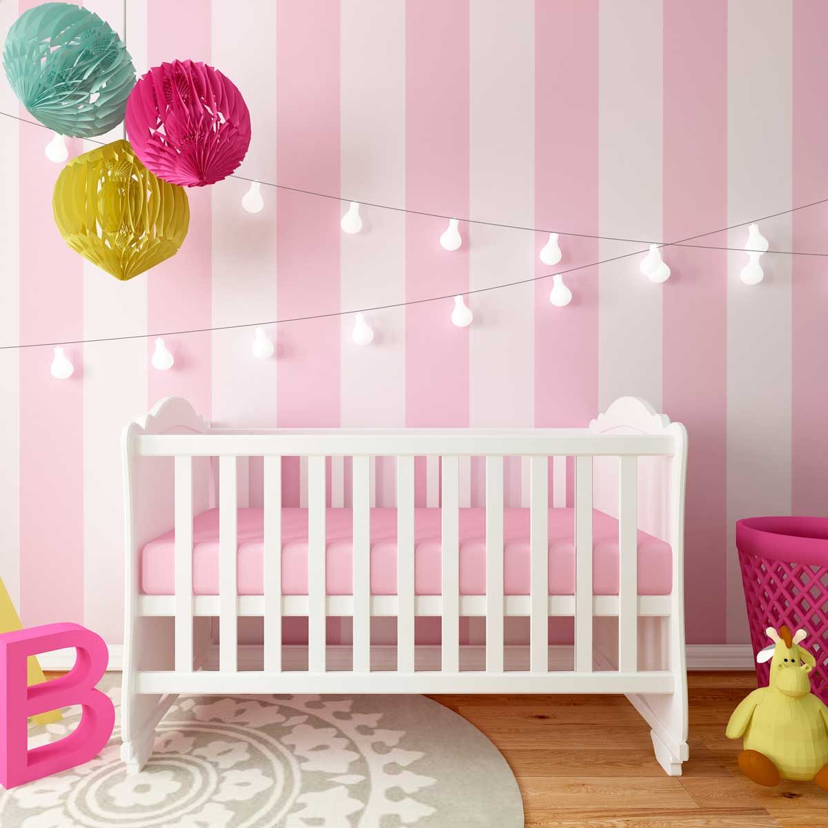 A white crib with a pink mattress rests on a patterned rug, surrounded by pastel decorations and a pink basket, within a softly lit pink-striped room.