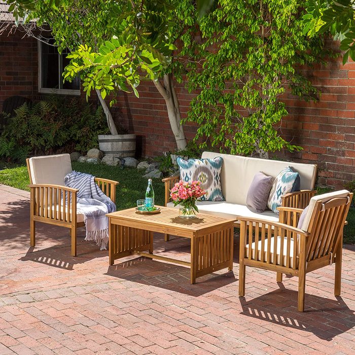 A wooden patio set features a sofa, two chairs, and a table with flowers, positioned on a brick surface under a leafy tree adjacent to a brick wall.