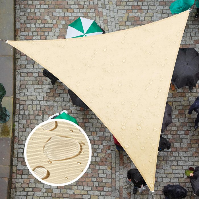 A large, triangular shade tarp collects rain droplets above a cobblestone area, while people with umbrellas walk below, sheltered from the weather.