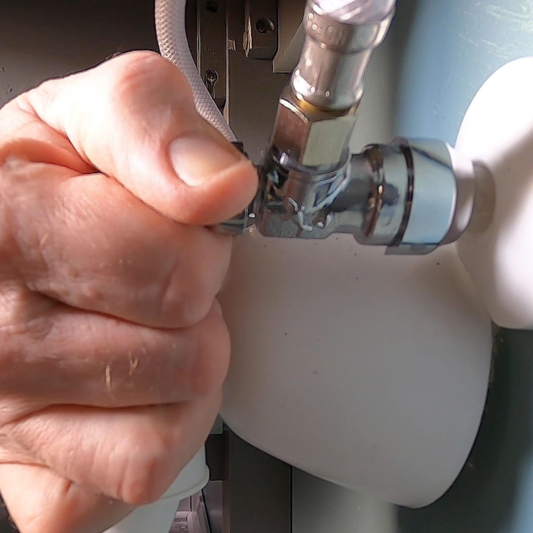 A hand grips a metallic valve, applying pressure to connect or disconnect it, with surrounding plumbing and machinery visible in a likely domestic or mechanical setting.