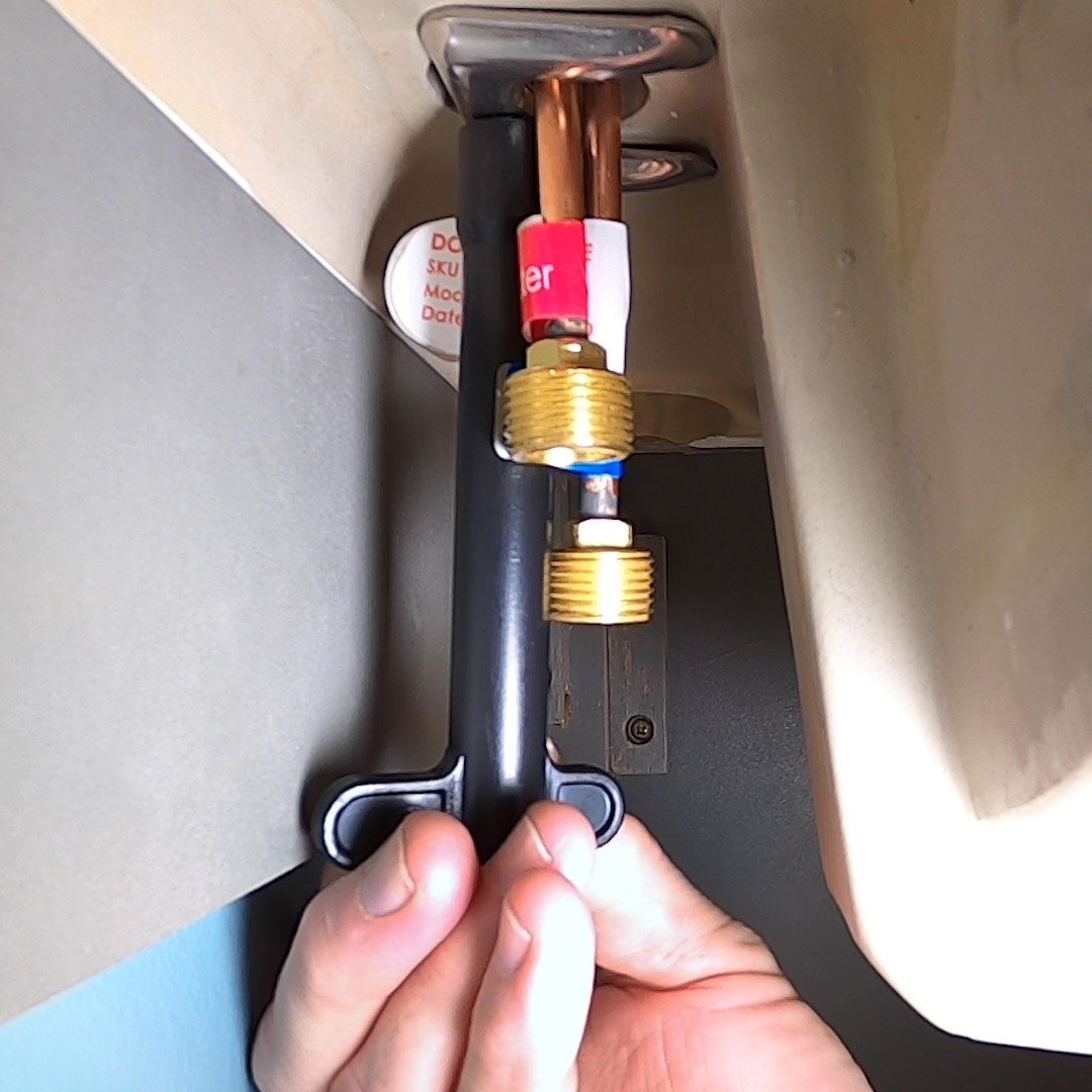 A hand grips a black tool attached to a brass valve and copper pipe under a sink, while a white and red label is visible nearby.
