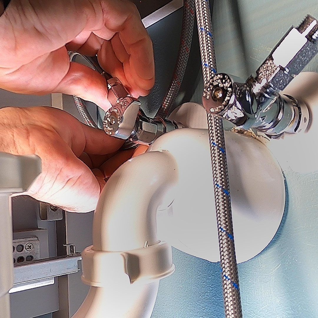 A hand adjusts a chrome plumbing valve connected to white piping, while flexible metallic hoses and a wall provide the surrounding context.