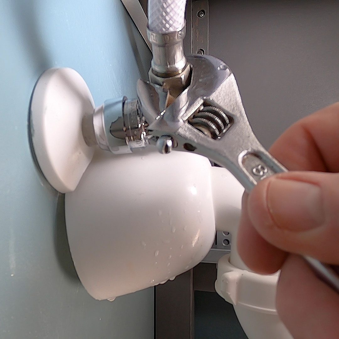 A hand grips a wrench, tightening a shiny metal fitting on a white pipe against a blue wall, with water droplets visible on the surface.