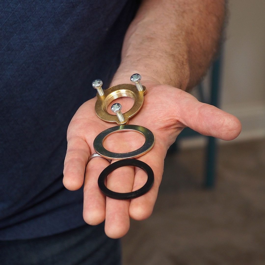 A hand holds four circular objects: a brass washer with screws on top, flanked by two flat washers, one metal and one black rubber.