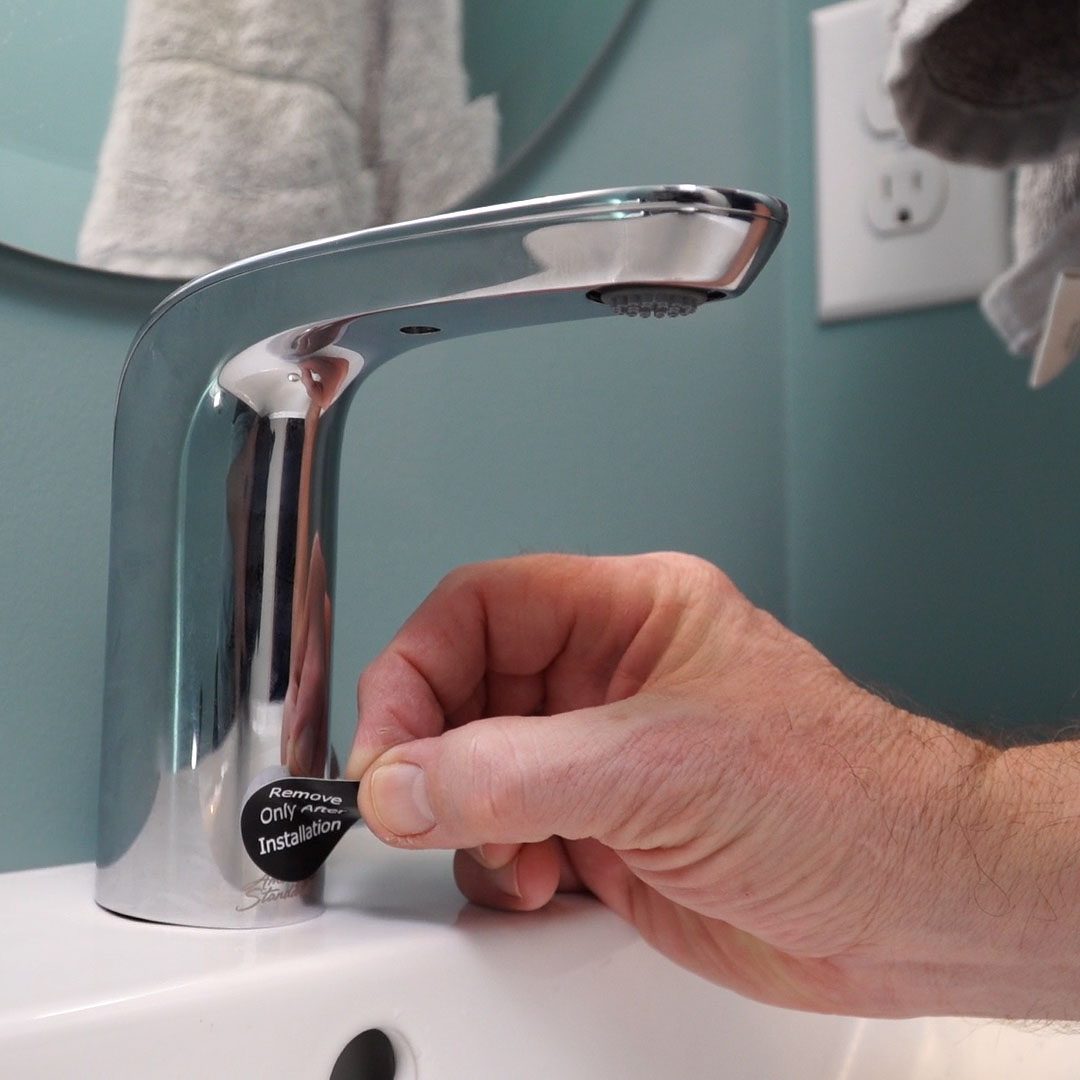 A hand removes a sticker from a polished bathroom faucet, indicating installation, against a backdrop of a blue wall and a round mirror.