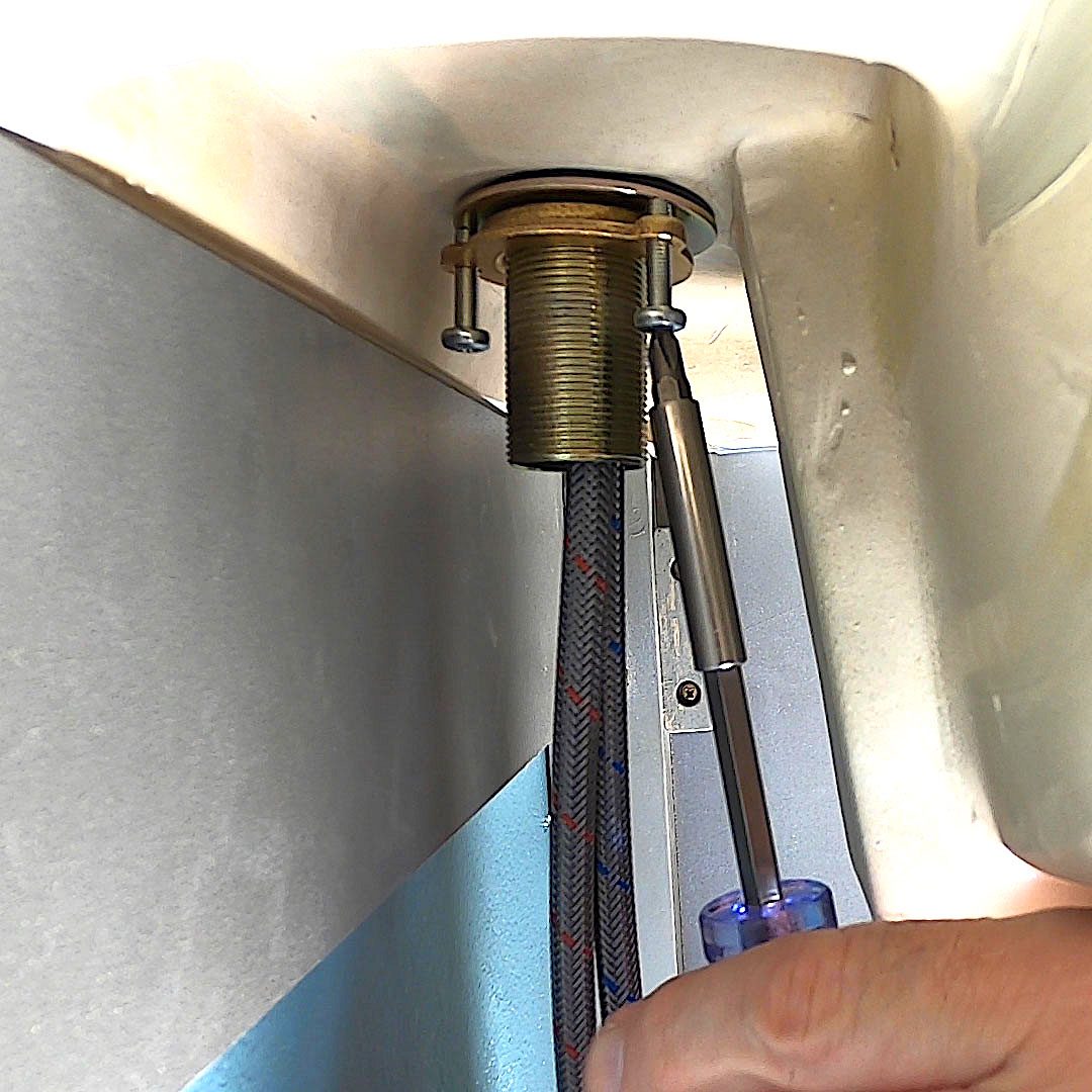 A hand uses a screwdriver to tighten a metal fitting connected to a hose, situated beneath a sink against a light-colored wall.