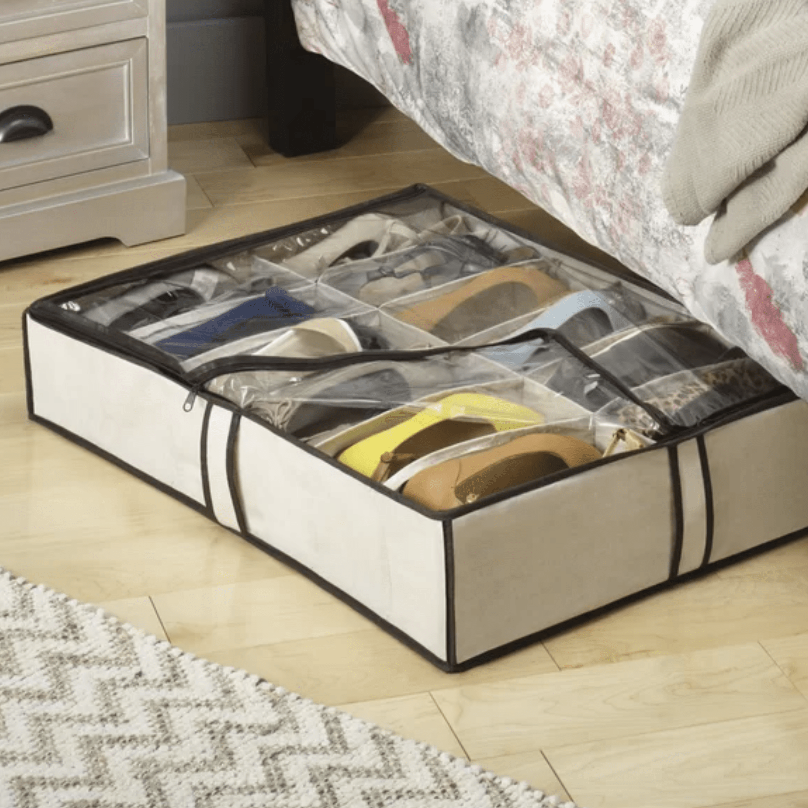 A storage container with a transparent lid holds multiple pairs of shoes, positioned under a bed in a bedroom with wooden flooring and a rug.