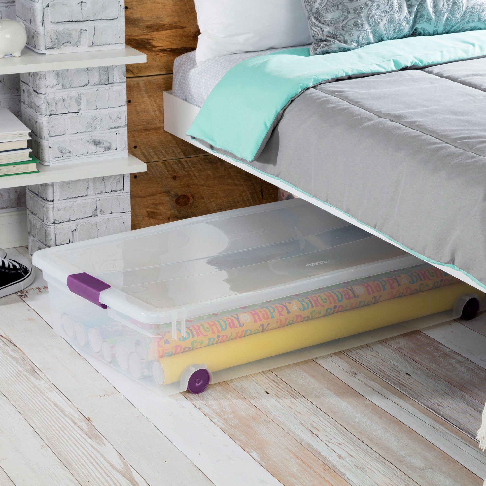 A clear storage bin holds rolled wrapping paper and is positioned under a bed in a cozy, modern bedroom with wooden accents and shelves.