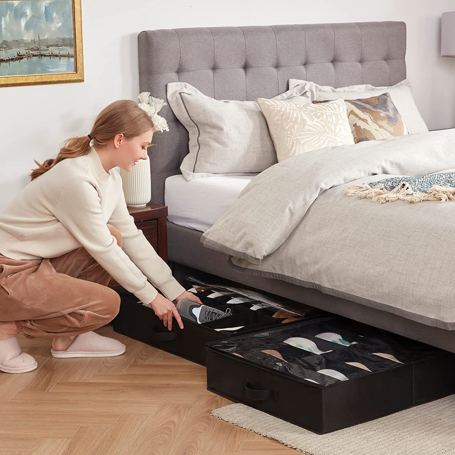 A woman crouches beside a bed, organizing shoe storage boxes placed under the frame in a cozy, well-decorated bedroom.