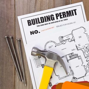 A hammer rests on a building permit and blueprint, while three nails lie beside it on a wooden surface.