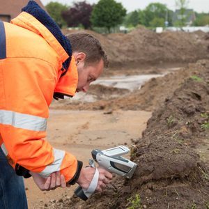 A man in an orange safety jacket examines soil with a handheld device in a construction area surrounded by dirt mounds and greenery in the background.