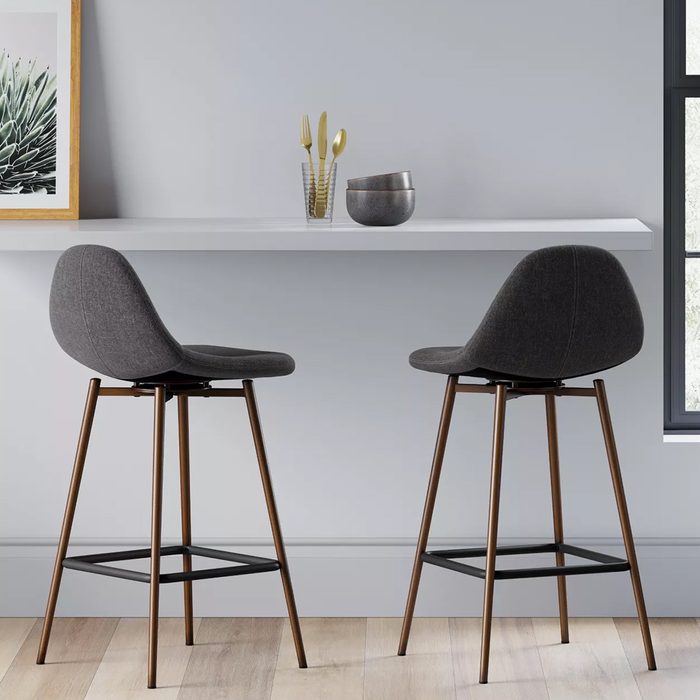 Two gray bar stools stand against a minimalist wall, beside a shelf displaying decorative items, including bowls and utensils, in a softly lit environment.
